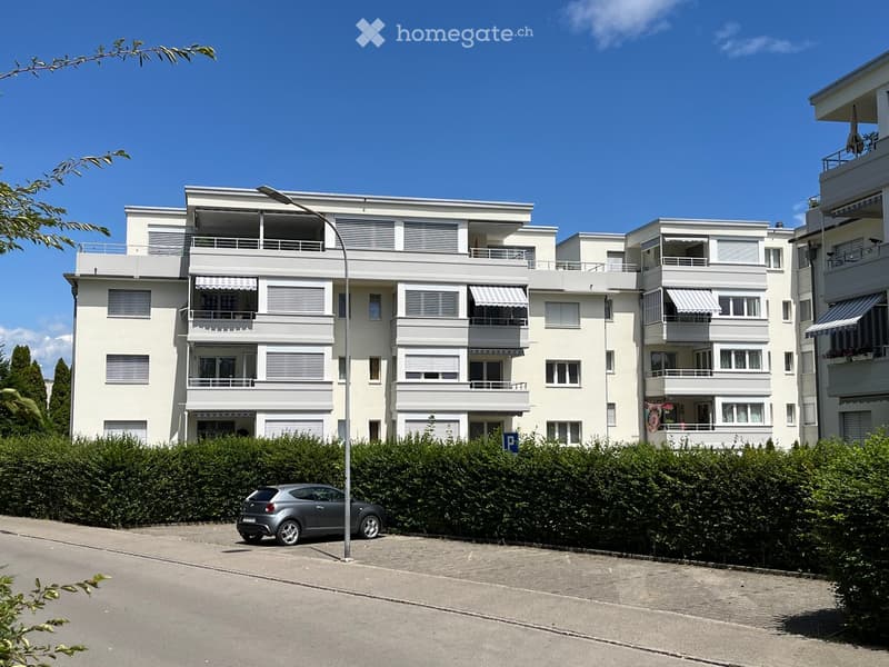 Modern white multi-story apartment building with balconies, awnings, and surrounding greenery.