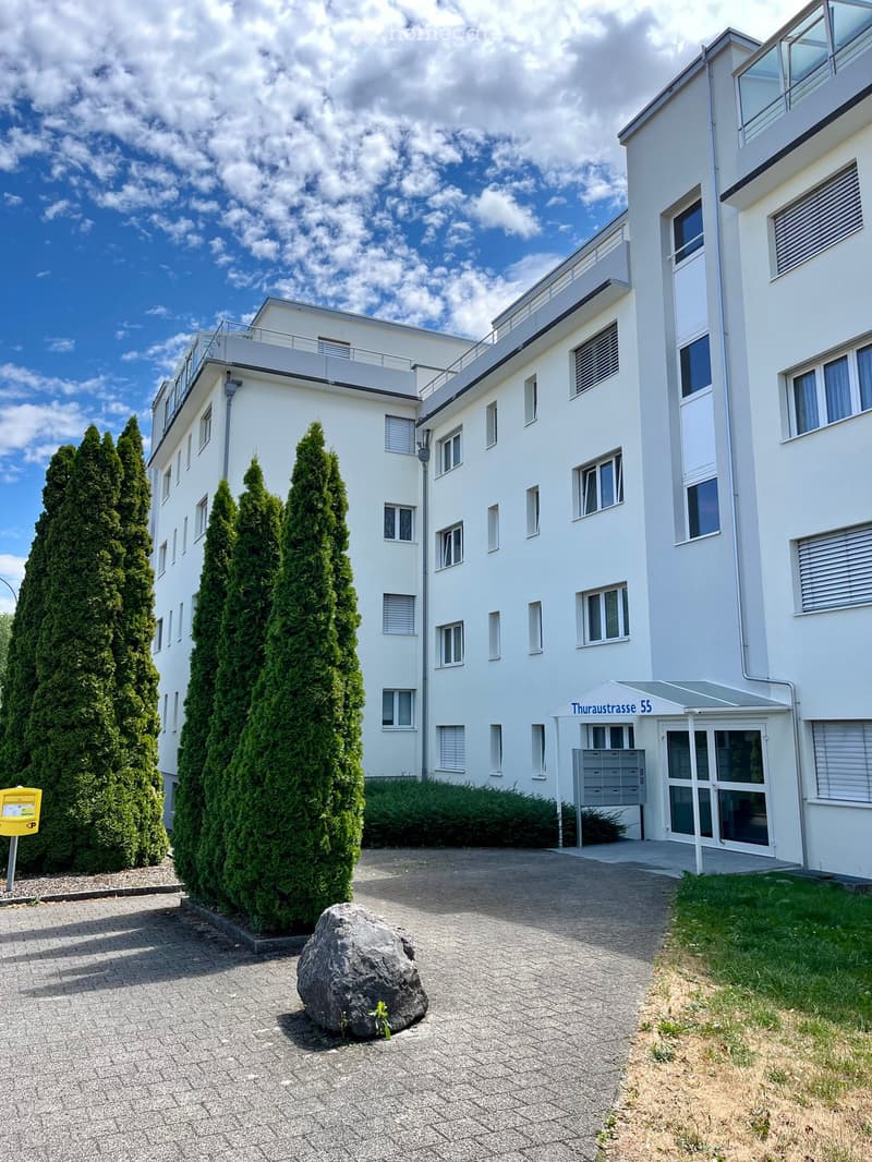 Modern white multi-story building with large windows, balconies, and a paved entrance area with greenery and a large rock.