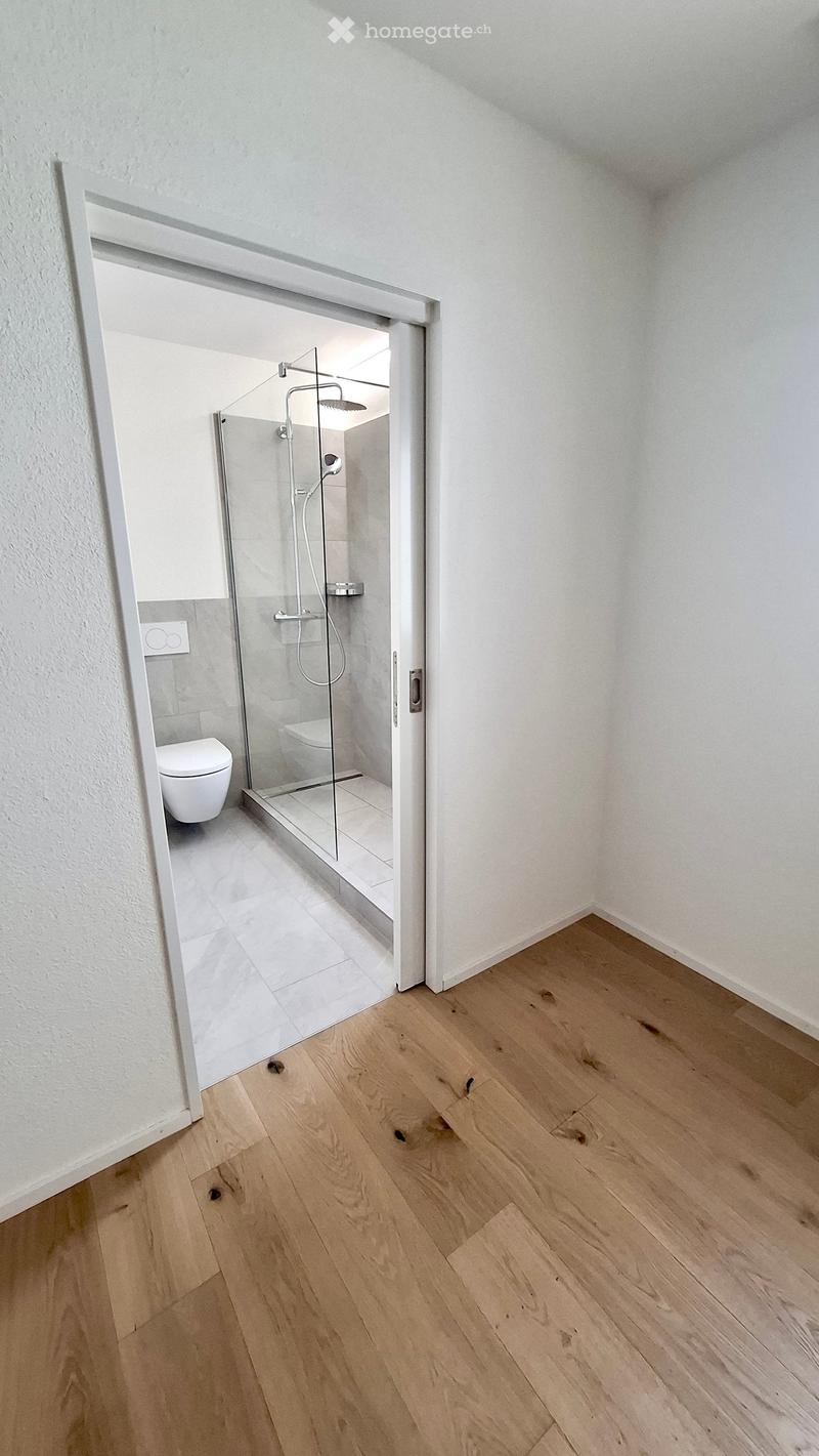 Empty room with light wooden flooring and white walls, featuring an open doorway leading to a modern bathroom with a glass-enclosed shower and a toilet.