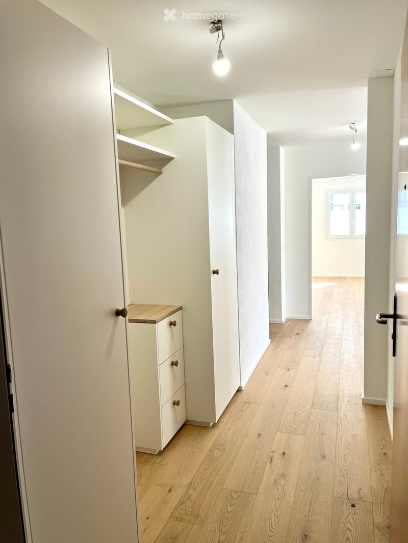 Empty hallway with light wooden flooring, white walls, built-in closet with shelves, and a small cabinet with drawers.