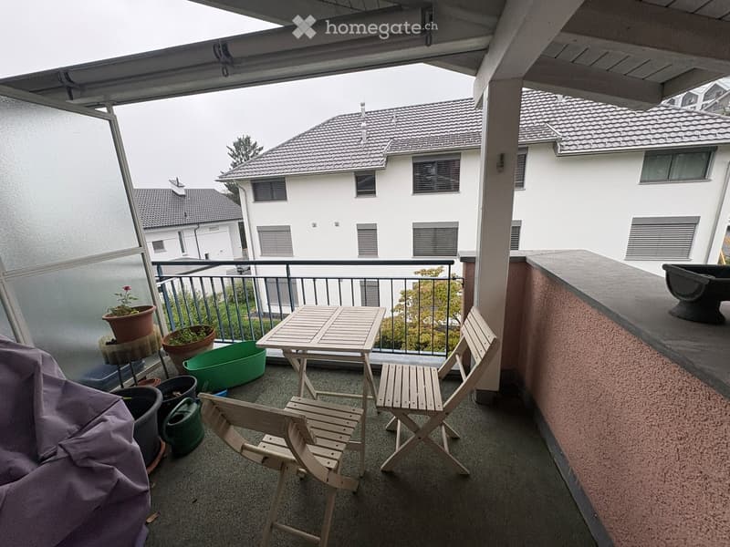 Balcony with a small table, two chairs, several potted plants, and a garden hose. The balcony has a railing and overlooks neighboring buildings.