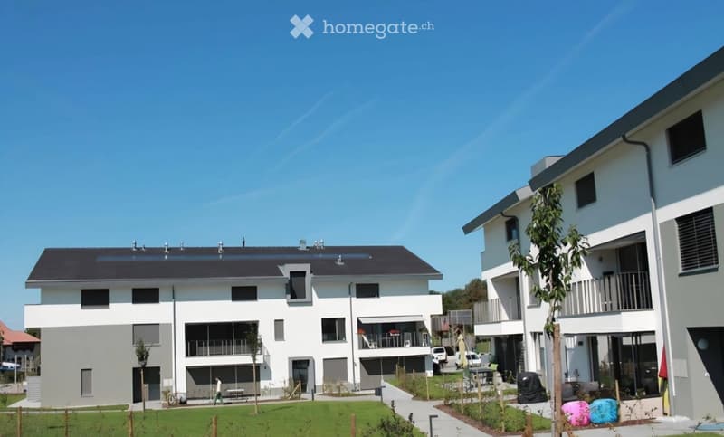 Modern two-story residential building with white exterior, large windows, and a balcony. The building features a flat roof and contemporary design, situated in a well-maintained garden area.