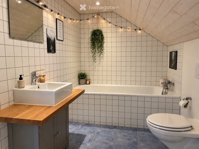 Modern bathroom with white tiles, a wooden vanity, a large sink, a bathtub, and a toilet. Features include a hanging plant, a mirror, and decorative candles.