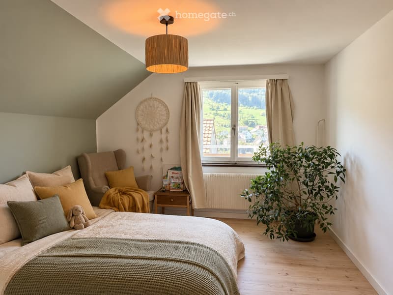 Cozy bedroom with a large window offering a scenic view, featuring a bed with pillows and a blanket, a sofa with cushions, a small table with books, a potted plant, and a dreamcatcher on the wall.