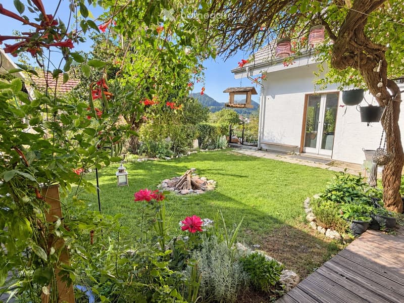 Well-maintained garden with a lawn, flower beds, a fire pit, and a bird feeder. A white house with large windows is visible in the background, along with a wooden deck and hanging planters.