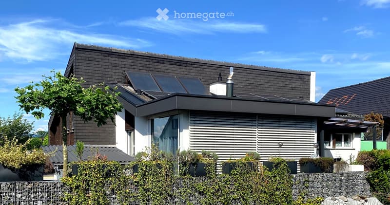 Modern residential building with a flat roof, solar panels, and a small outdoor seating area. The structure features large windows and a sleek, contemporary design.