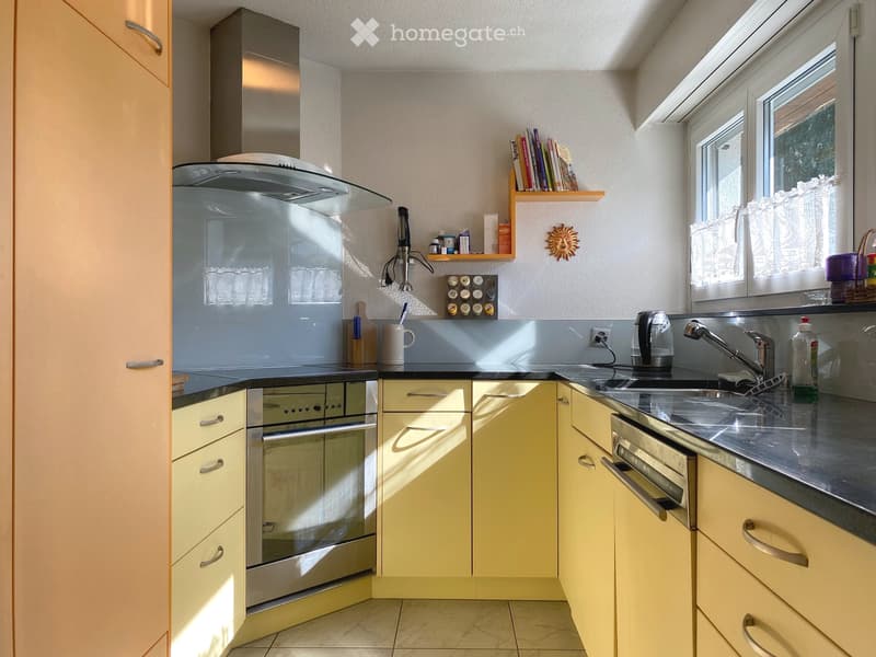 Modern kitchen with light-colored cabinets, stainless steel appliances, and a small window with a curtain. Includes a faucet, oven, microwave, refrigerator, and a small bookshelf with books.