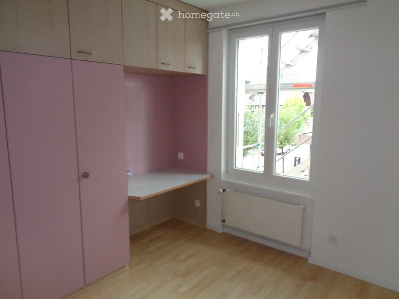 Empty room with light pink walls, wooden floor, built-in cabinets, and a window with a view of the street.