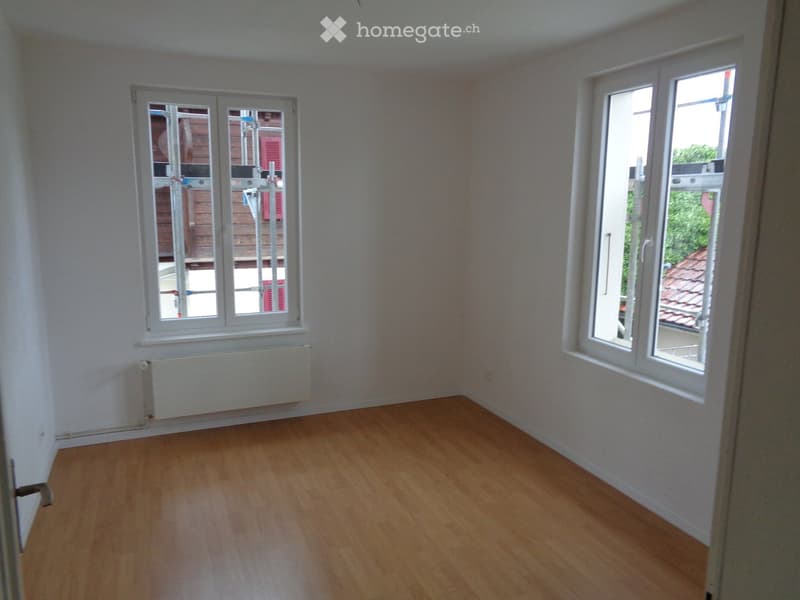 Empty room with wooden flooring, white walls, and two windows with white frames. A radiator is visible under one window.