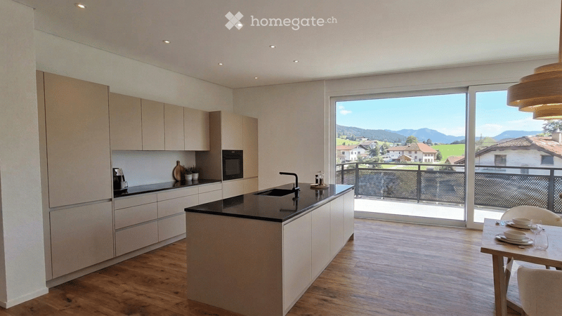 Modern kitchen with light wood cabinets, black countertop, island, and a view of the mountains through large windows and a balcony.