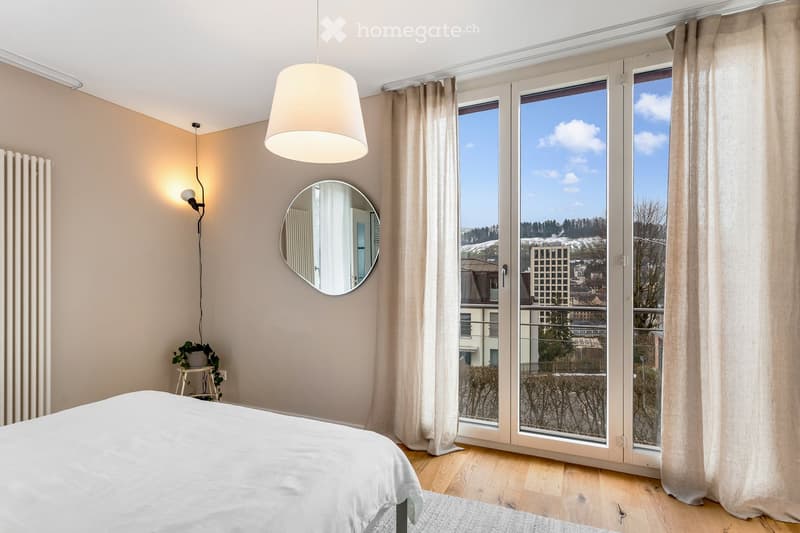 A bedroom with a bed, round mirror, wall-mounted lamp, and large windows with a view of the cityscape.