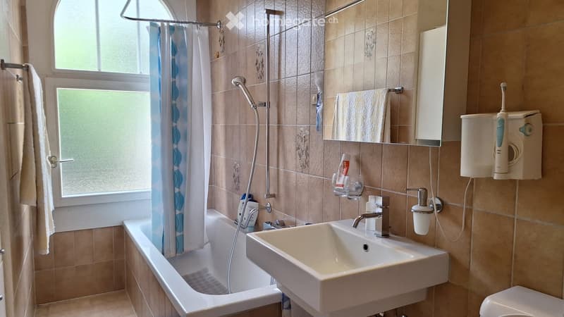 Bathroom with a bathtub, sink, and toilet. Features a showerhead with a blue and white shower curtain, a towel on a rack, a mirror, and a toothbrush holder.