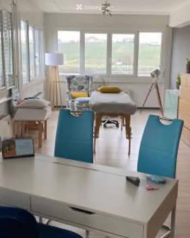 Brightly lit room with large windows, a massage table, chairs, a sofa, and a lamp. The space is well-organized and appears to be used for relaxation or therapy.