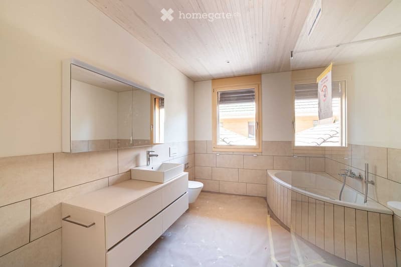 Modern bathroom with a white sink, bathtub, and large mirror. The room features light-colored tiles and wooden accents.