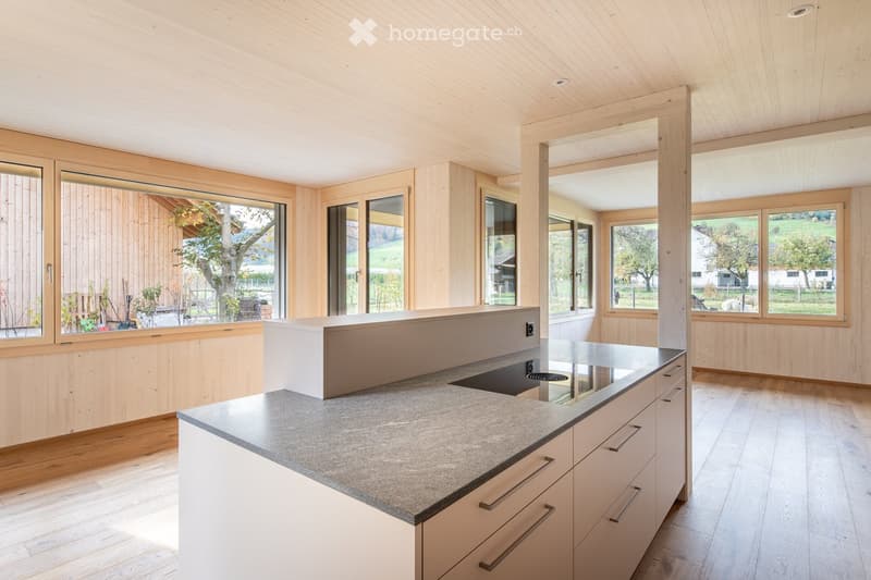 Modern kitchen with light wood paneling, white cabinets, and a grey countertop, featuring large windows overlooking a green landscape.