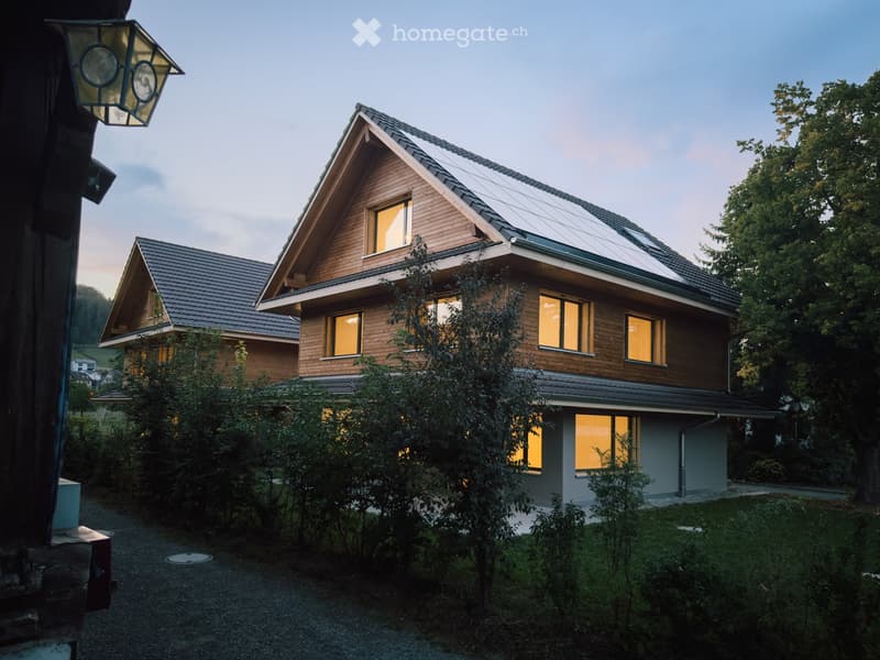 Two-story wooden house with dark gray roof, large windows, and solar panels on the roof, surrounded by greenery.