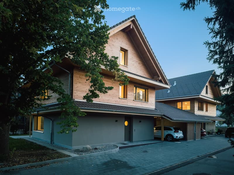 Two-story modern house with wooden facade, grey garage, and multiple windows. Features a driveway with parked cars and surrounding greenery.