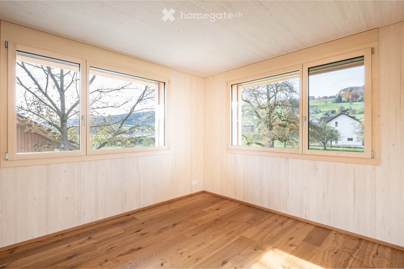 Empty room with light wooden walls, wooden floor, and large windows offering a scenic view of trees and countryside.