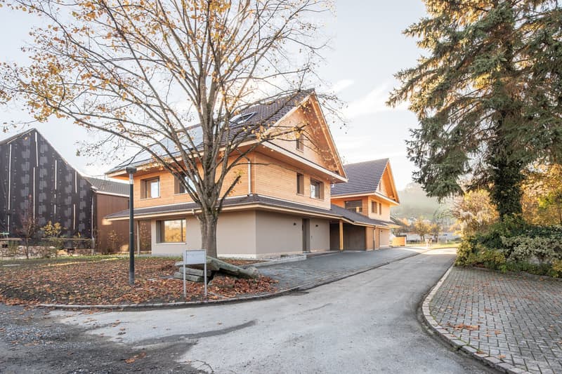 Two-story modern house with wooden facade, large windows, and a sloped roof. The house features a driveway and is surrounded by trees and greenery.