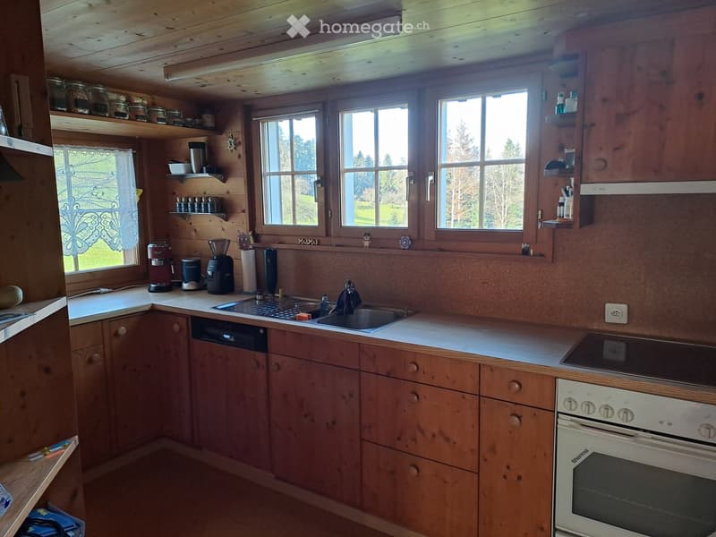 Cozy wooden kitchen with large windows, sink, oven, dishwasher, coffee maker, blender, and shelves with jars and bottles.