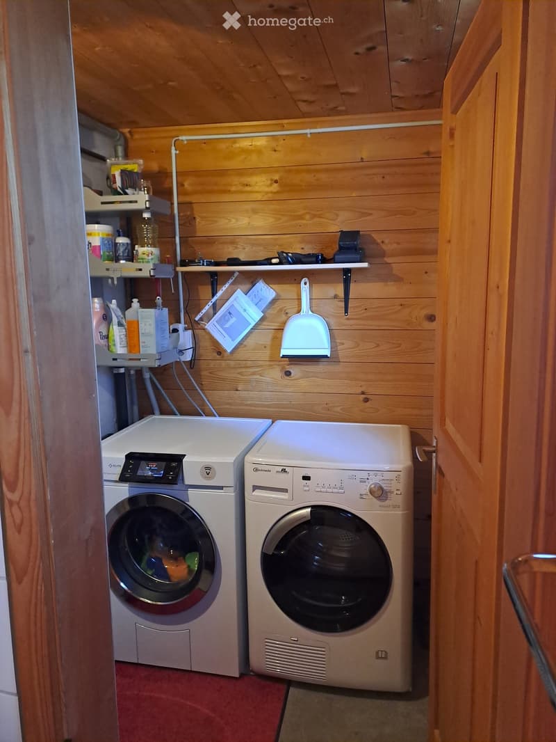 A small, wooden-paneled laundry area with a washing machine and dryer side by side, shelves holding cleaning supplies, and a broom hanging on the wall.