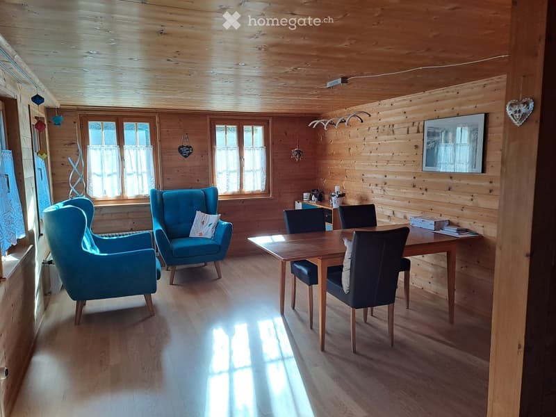 Cozy living room with wooden walls and ceiling, large windows, blue armchairs, wooden dining table with chairs, and a mirror on the wall.