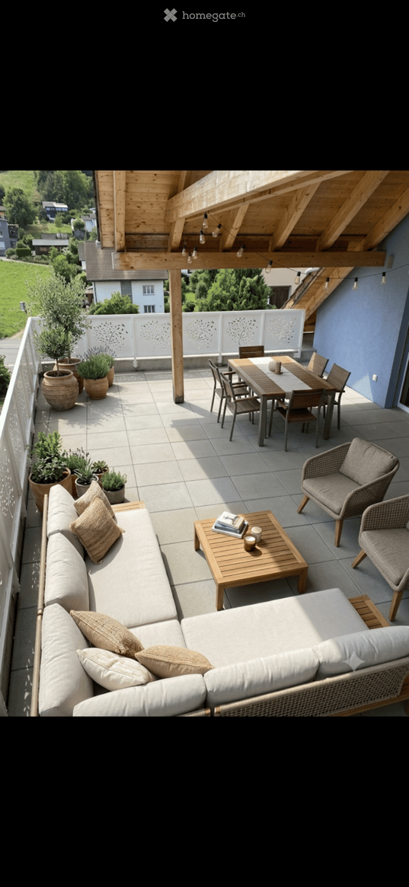 A spacious, well-lit balcony or terrace with a wooden pergola, outdoor seating including a sofa, coffee table, and dining set, surrounded by potted plants and offering a scenic view of a green landscape.