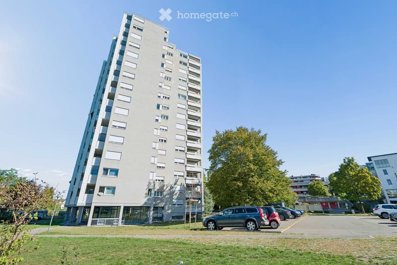 Modern multi-story apartment building with balconies, surrounded by trees and a parking lot with several cars.