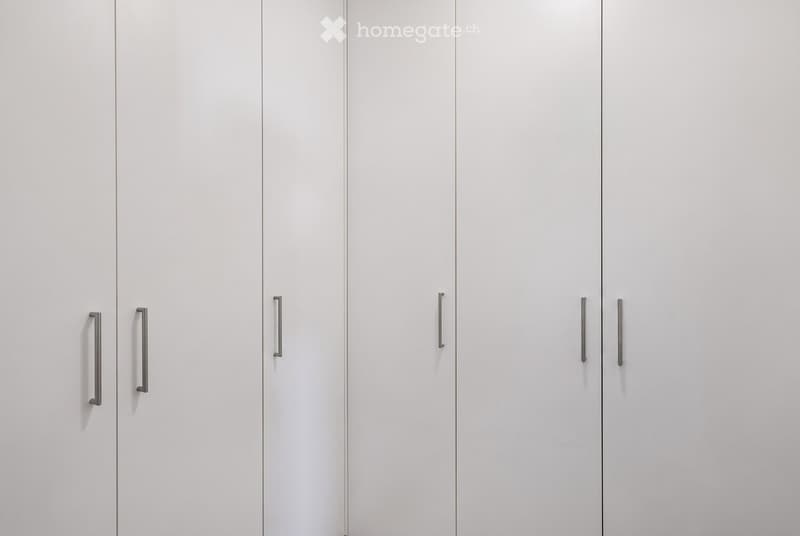 A completely empty room with white walls and white built-in wardrobes with silver handles.