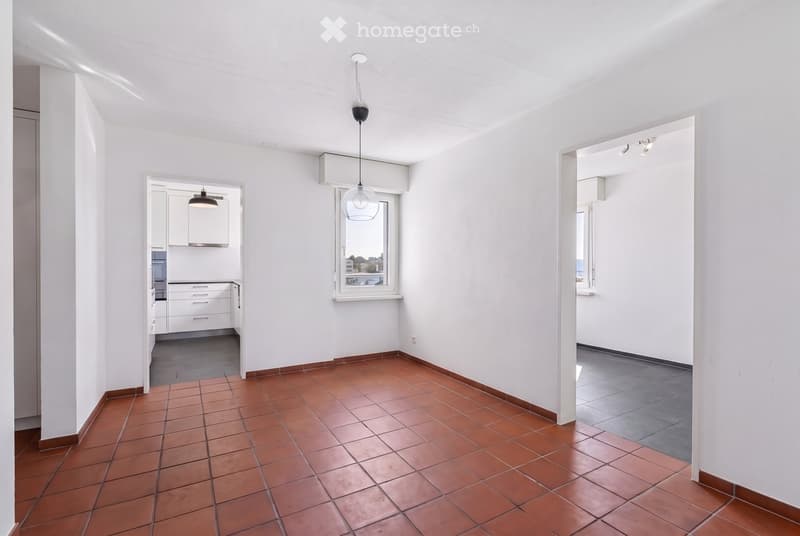 Empty room with white walls, red tiled floor, and a view of a kitchen area through an open doorway. The space is unfurnished with no visible furniture or equipment.