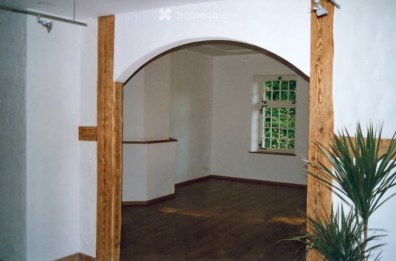 Empty room with wooden floor, white walls, wooden pillars, arched doorway, small window, and a plant.