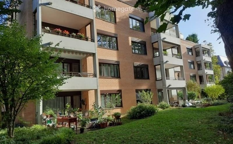 Multi-story apartment building with brick facade, multiple balconies, and greenery in the foreground.