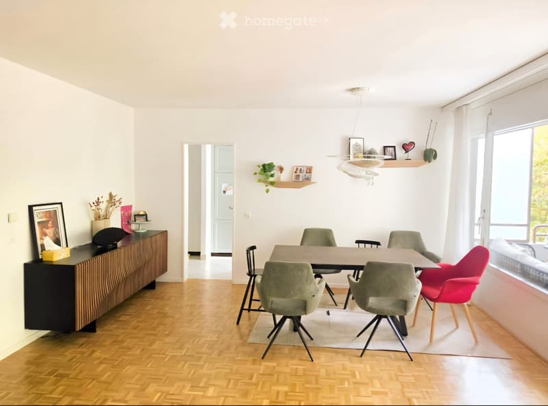 Modern dining area with wooden floor, dining table surrounded by chairs, cabinet with picture frames and decorative items, and a hanging lamp.