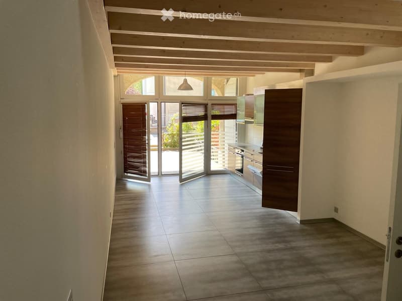 Bright hallway with tiled flooring, wooden ceiling beams, and large glass doors leading to an outdoor area. The hallway connects to a modern kitchen with stainless steel appliances.