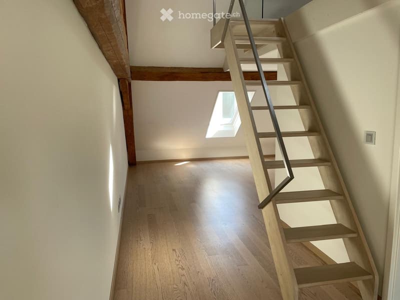 Spacious hallway with wooden flooring, exposed wooden beams, and a modern staircase with a metal railing leading upwards. Natural light streams in from a skylight at the top of the stairs.