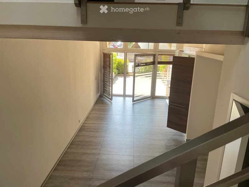 Bright hallway with tiled floor, white walls, and large glass doors leading to an outdoor area. Wooden railing visible on the staircase.