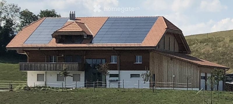 Modern two-story house with a wooden facade, white lower section, and solar panels on the roof. Features a balcony on the upper level and is situated in a green, hilly landscape.