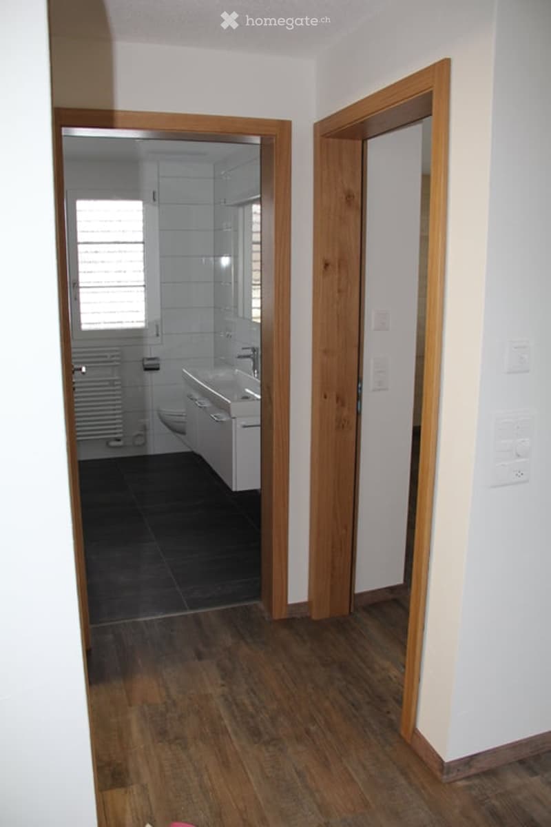 A hallway with wooden flooring and white walls, leading to a bathroom with white tiles, a sink, and a toilet.