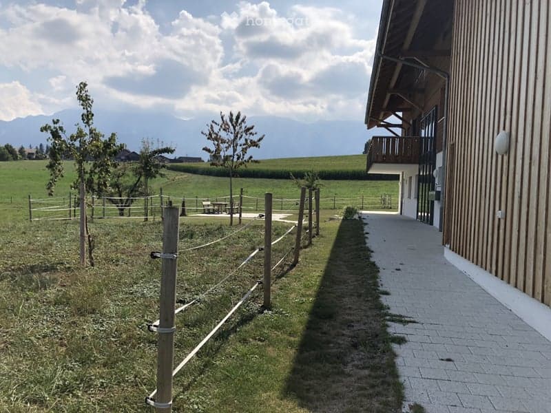 Outdoor area with a fenced lawn, a few trees, a pathway leading to a building, and a scenic mountain view in the background.