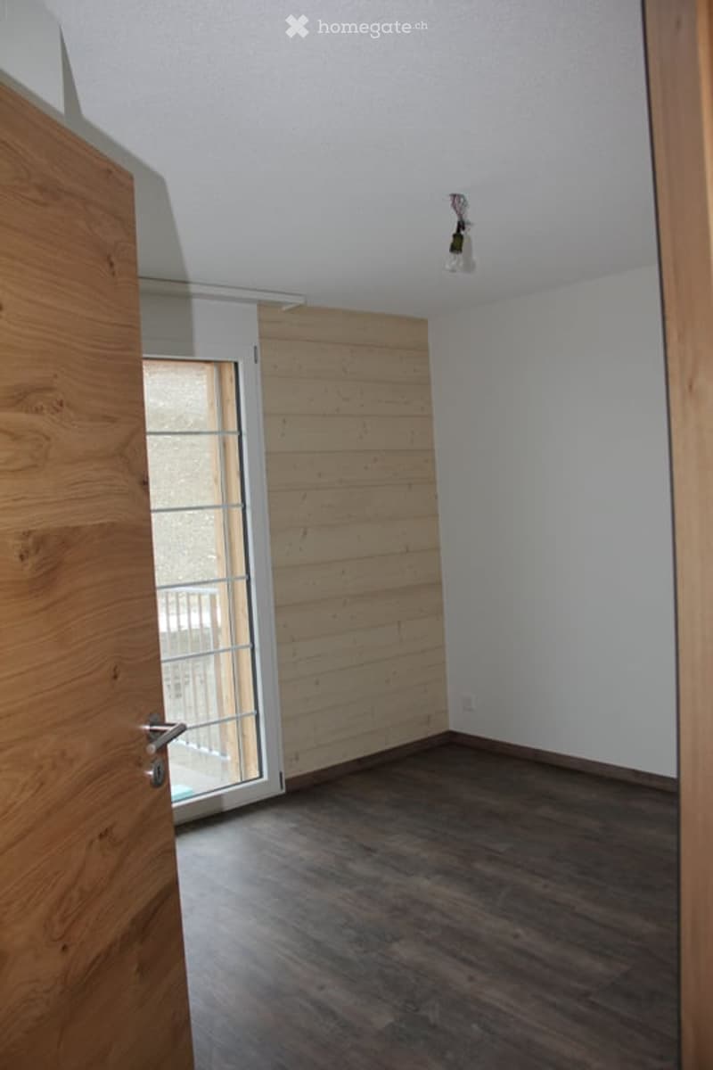 Empty room with wooden flooring, white walls, and a large window with a view of a balcony. No furniture or equipment present.