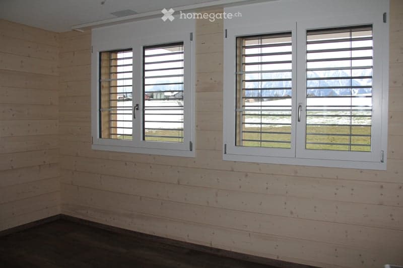 Empty room with wooden walls and flooring, featuring two large windows with white shutters overlooking a snowy landscape.