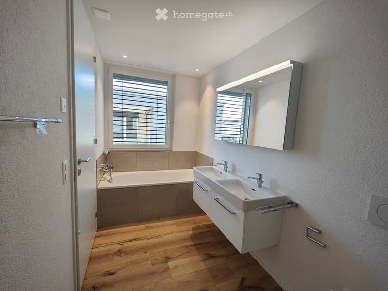 Modern bathroom with a bathtub, double sink, large mirror, and wooden flooring.