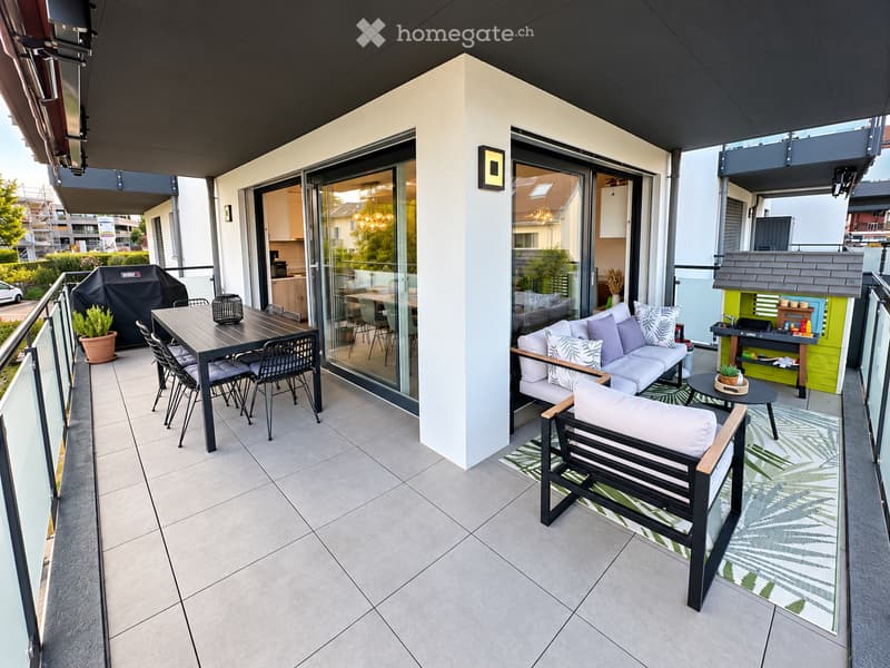 Modern balcony with grey sofa, white cushions, black bench, dining table, chairs, and a small grill. Features a green rug and potted plants.