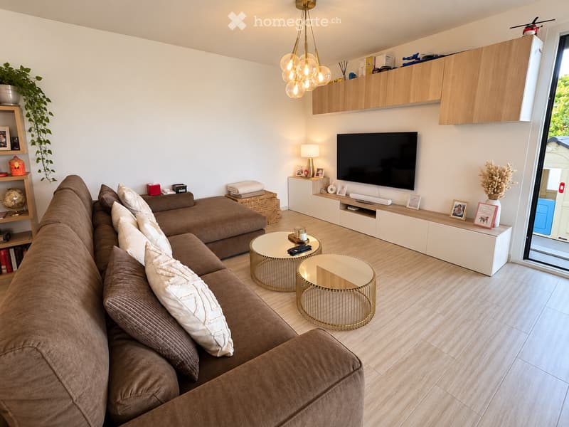 Modern living room with a brown sectional sofa, two round gold-framed coffee tables, a flat-screen TV on a white console, a hanging chandelier, and decorative shelves with plants and picture frames.