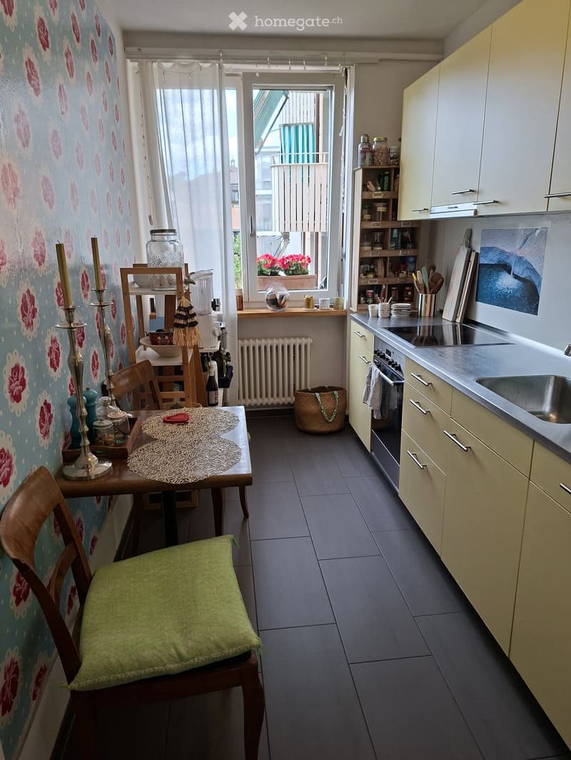 A small, well-lit kitchen and dining area with a table, chairs, and various kitchen appliances. The space features a mix of modern and vintage decor, including a floral wallpapered wall, a small dining table with a lace table runner, and a wooden chair wi