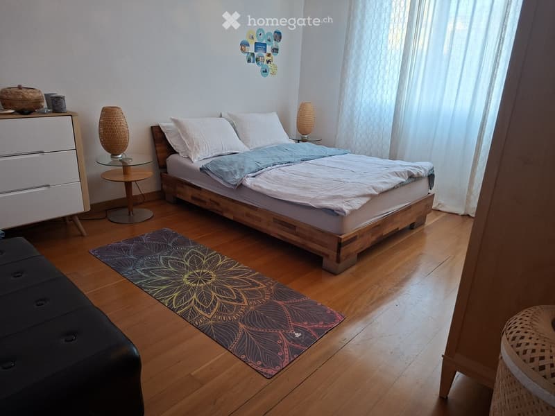 Cozy bedroom with wooden floor, bed with white linens, nightstand with lamp, side table, and decorative rug.