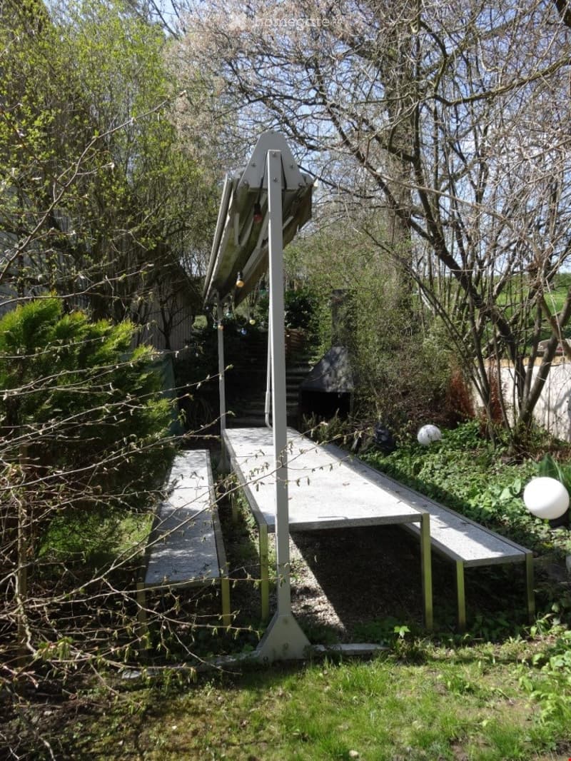 Outdoor area with a modern, elevated platform structure surrounded by greenery and trees. The platform includes a flat surface and steps leading up to it, with a small canopy or shelter on one side.
