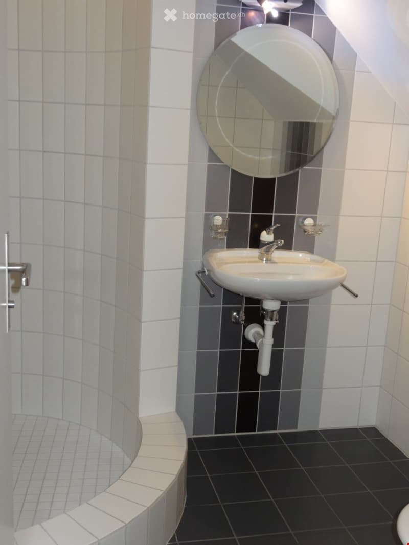 Modern bathroom with white tiled walls, black tiled floor, a white sink with a chrome faucet, a round mirror, and a soap dispenser.