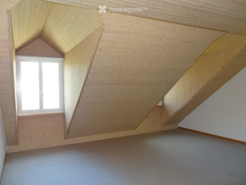 Empty attic room with wooden walls and ceiling, grey carpeted floor, and a single window letting in natural light.
