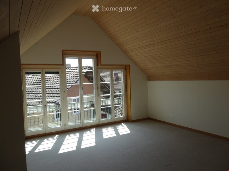 Empty attic room with wooden ceiling, large windows, and carpeted floor. No furniture or equipment present.